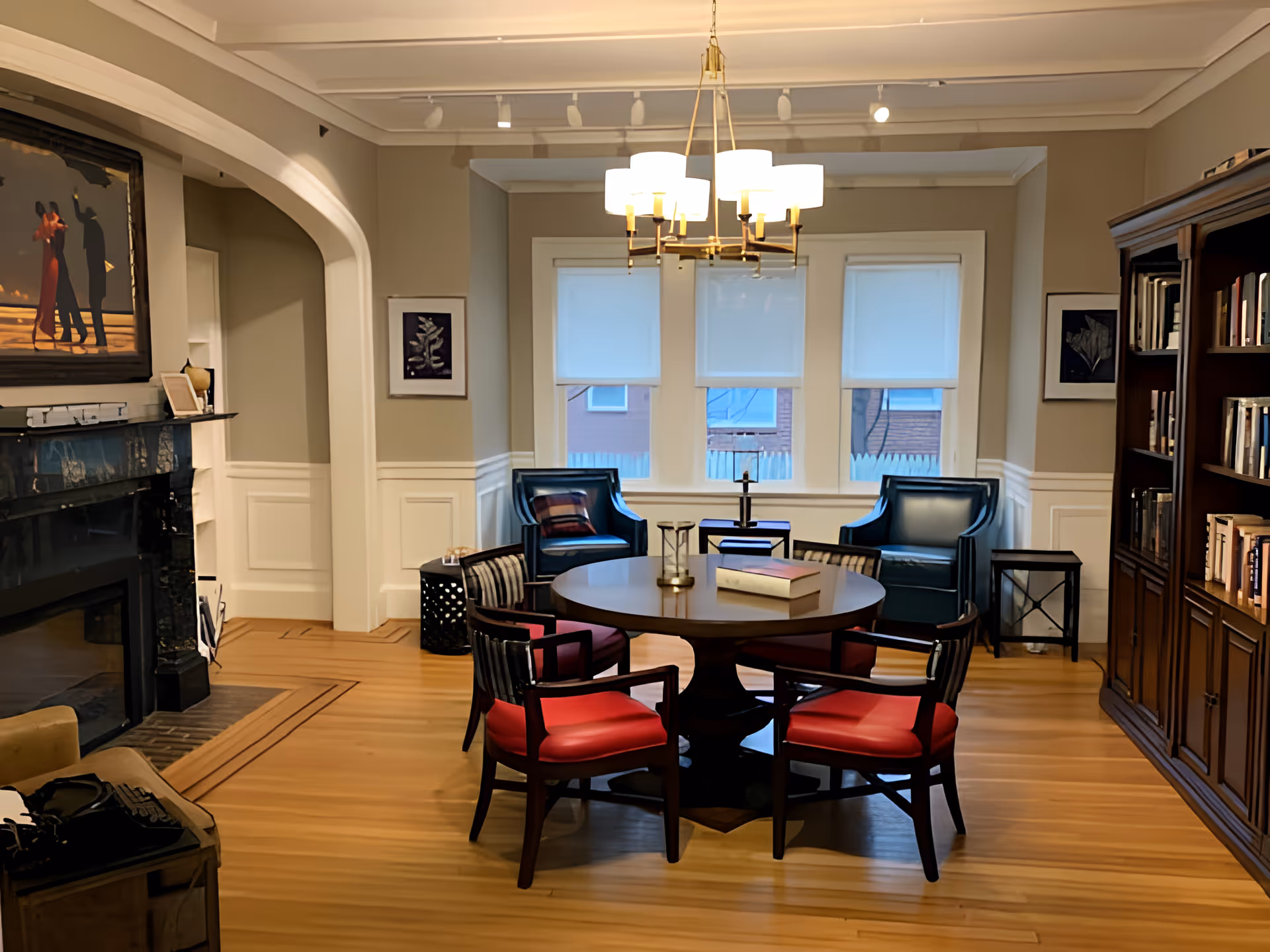 A cozy living room with a round wooden table surrounded by four chairs with red cushions. Behind the table are two blue armchairs placed in front of three windows with white blinds. A chandelier with multiple lights hangs from the ceiling. To the right is a large wooden bookshelf filled with books, and to the left is a black marble fireplace with a painting above it. The room has wooden flooring and beige walls with white wainscoting.