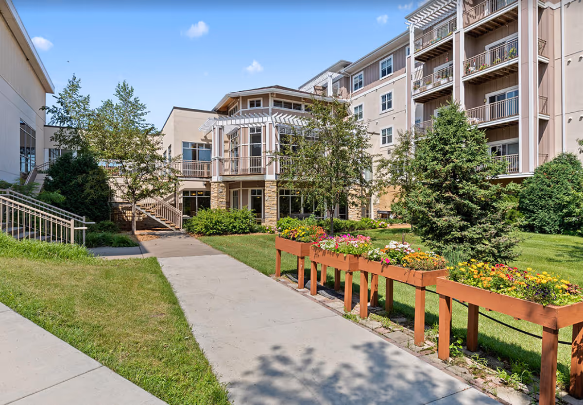 Outdoor view of McKenna Crossing senior living facility showing a paved walkway, green lawn, flower beds with colorful flowers, trees, and a multi-story building with balconies and large windows under a blue sky with a few clouds.