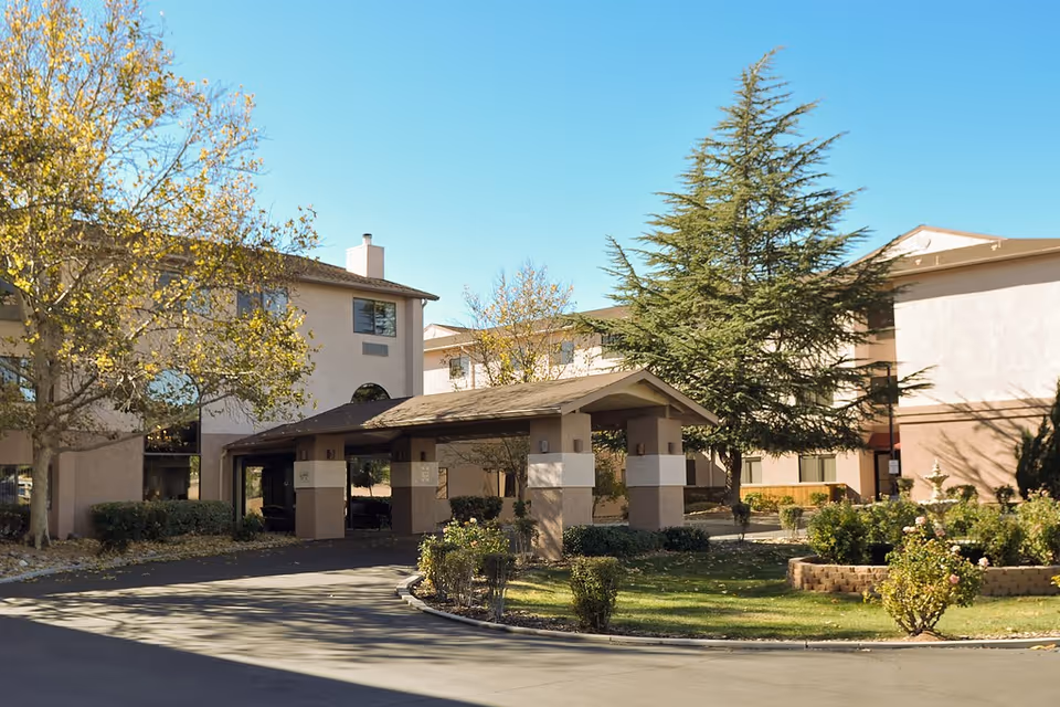 Exterior view of Cottonwood Village senior living facility showing a covered entrance with columns, surrounded by trees and landscaped greenery under a clear blue sky.