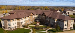 Aerial view of a large senior living facility building with multiple wings, brown roofs, beige and stone exterior walls, surrounded by green lawns and pathways, with trees and a partly cloudy sky in the background.