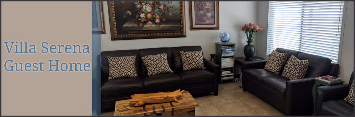 A cozy living room with two dark leather sofas adorned with patterned cushions, a wooden coffee table, a side table with a vase of flowers, a small bookshelf, and framed artwork on the walls. A window with blinds allows natural light into the room. The text 'Villa Serena Guest Home' is displayed on the left side of the image.
