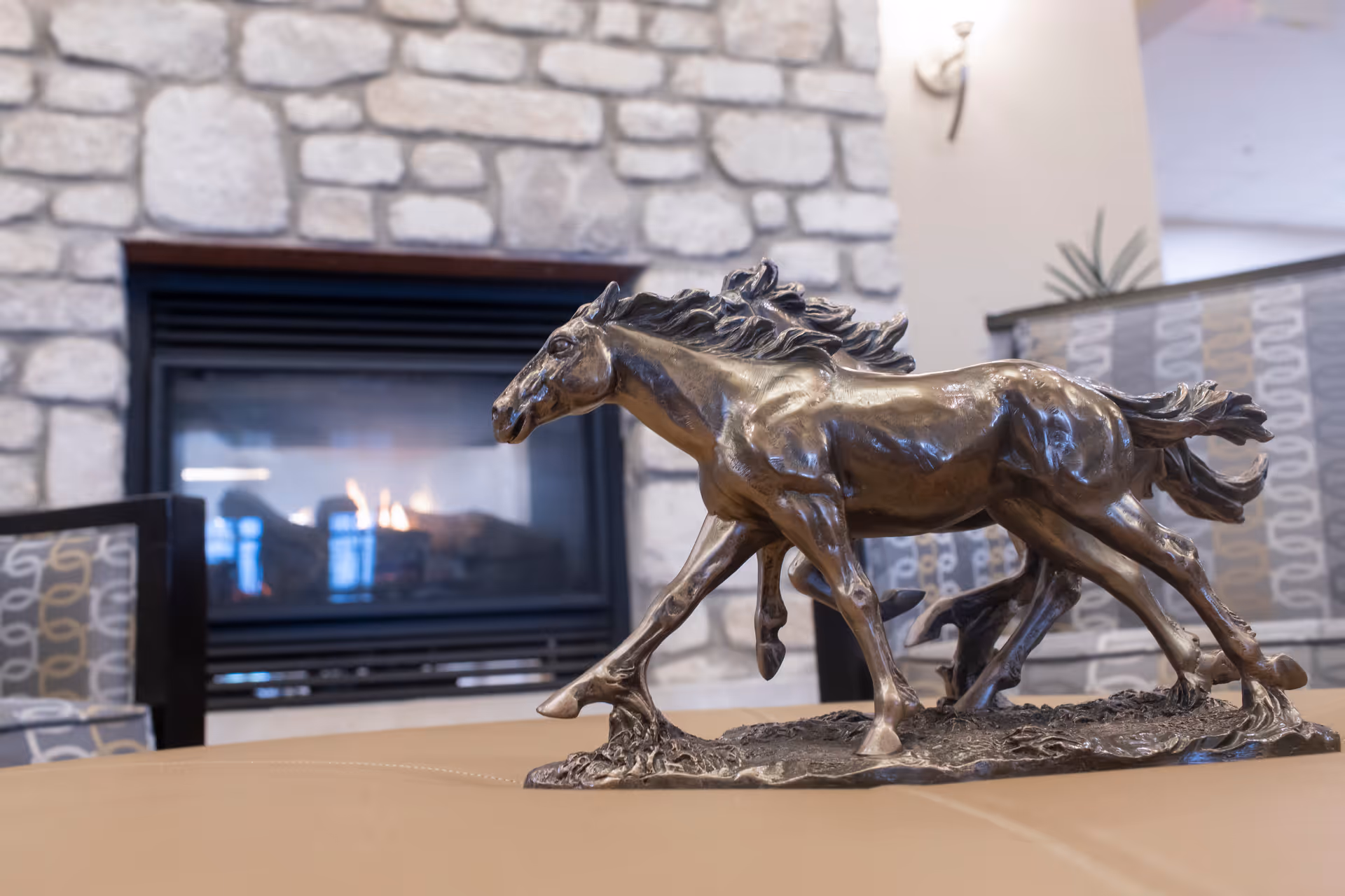 A bronze horse sculpture placed on a table in front of a stone fireplace with a fire burning inside. The background includes patterned chairs and a softly lit interior space.