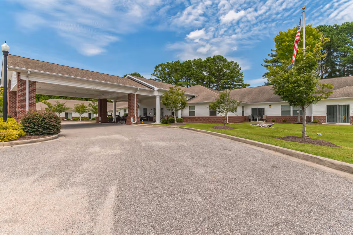 Front entrance of a single-story senior living facility with a covered porte-cochère, driveway, flagpole, and landscaped grounds.
