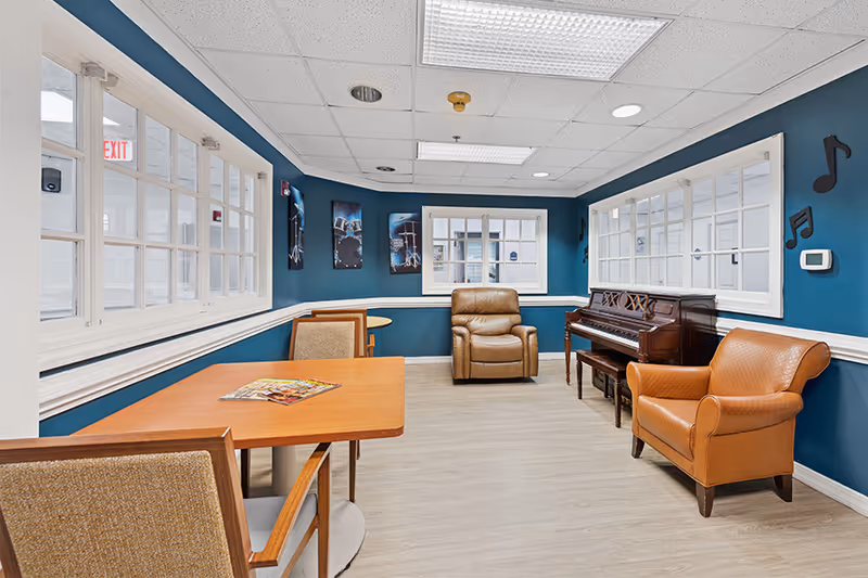 A cozy room with blue walls featuring musical notes decorations, a wooden piano, two brown leather armchairs, a wooden table with chairs, and framed pictures on the wall. The room has large windows with white frames and light-colored flooring.
