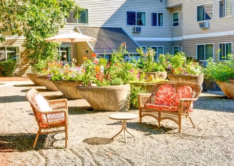 Outdoor courtyard with wicker chairs and a small table near large flower-filled concrete planters in front of a multi-story building.