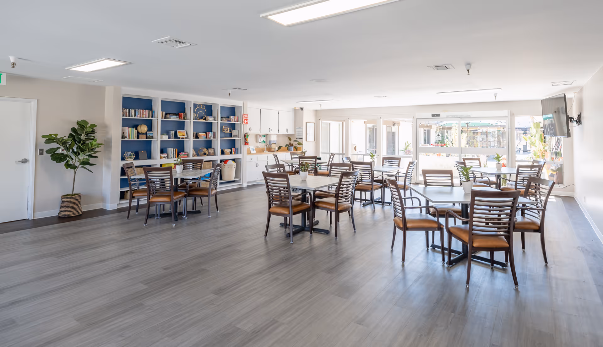 A bright and spacious dining area with multiple tables and chairs arranged neatly. There is a large bookshelf filled with books and decorative items against one wall, and large windows letting in natural light. The floor is covered with light-colored wood flooring, and there are plants and a television mounted on the wall.