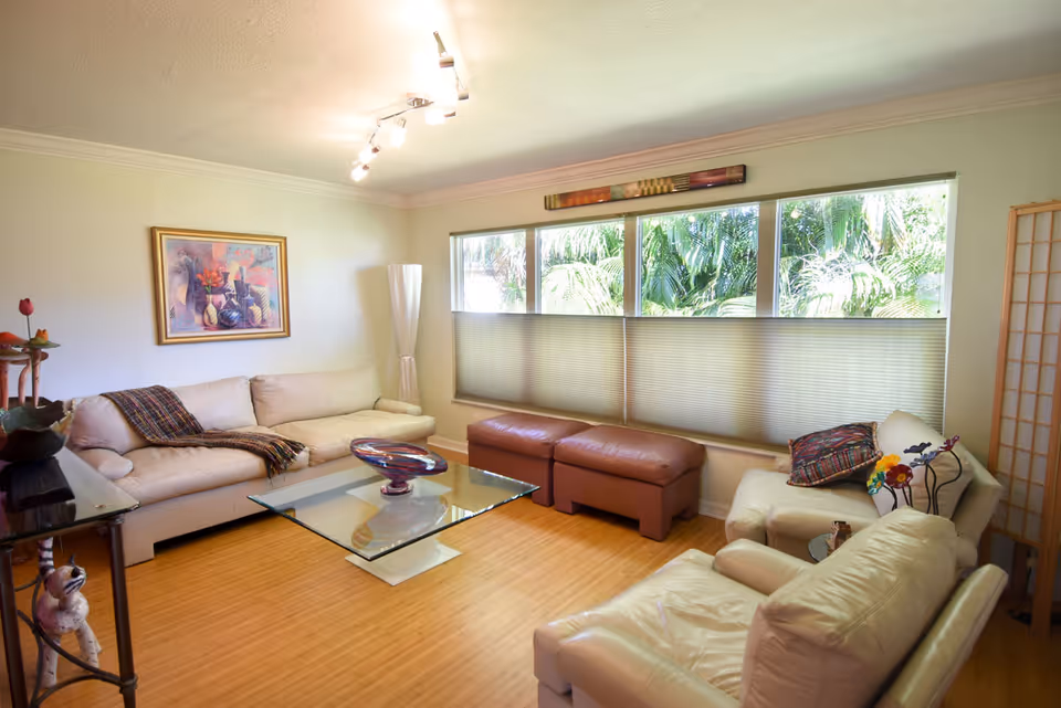 Bright living room with cream sofas, a glass coffee table, brown ottomans, and large windows showing tropical foliage.