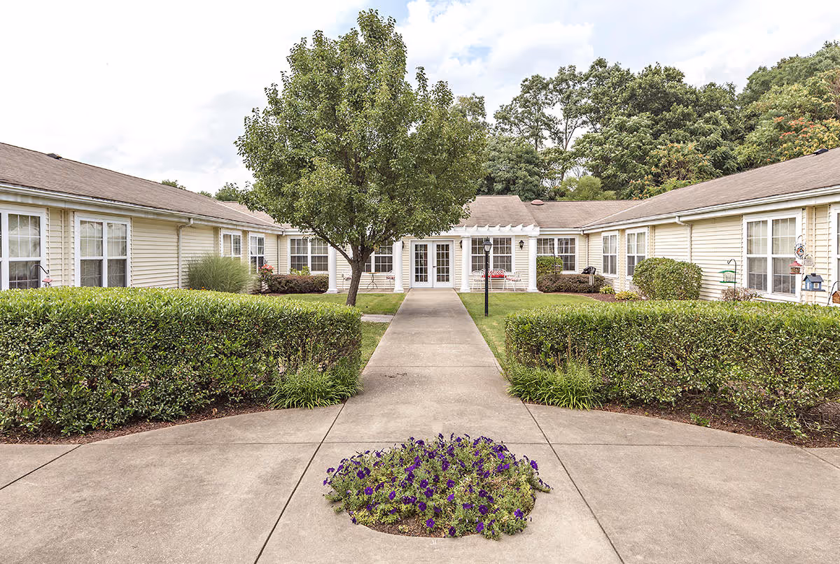 Outdoor view of The Addison of Logan Place showing a courtyard with a concrete walkway leading to a building entrance. The courtyard features trimmed hedges, a tree in the center, and a small flower bed with purple flowers. The building has light-colored siding and multiple windows.