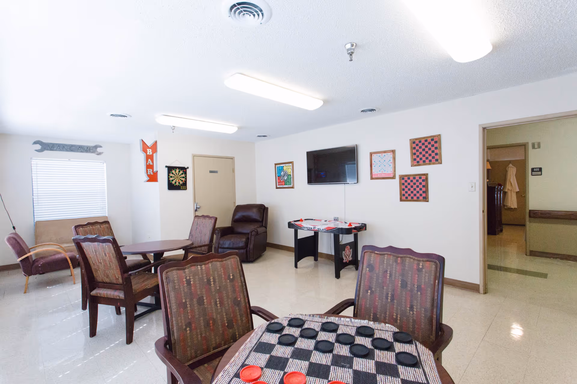A recreational room with several chairs and tables, including a table with a checkers board and pieces in the foreground. The room has a dartboard on the wall, a mounted flat-screen TV, and an air hockey table beneath it. There are framed checkerboards and other artwork on the walls, a brown recliner chair, and a window with blinds. The room is brightly lit with ceiling lights and has a door leading to a hallway.