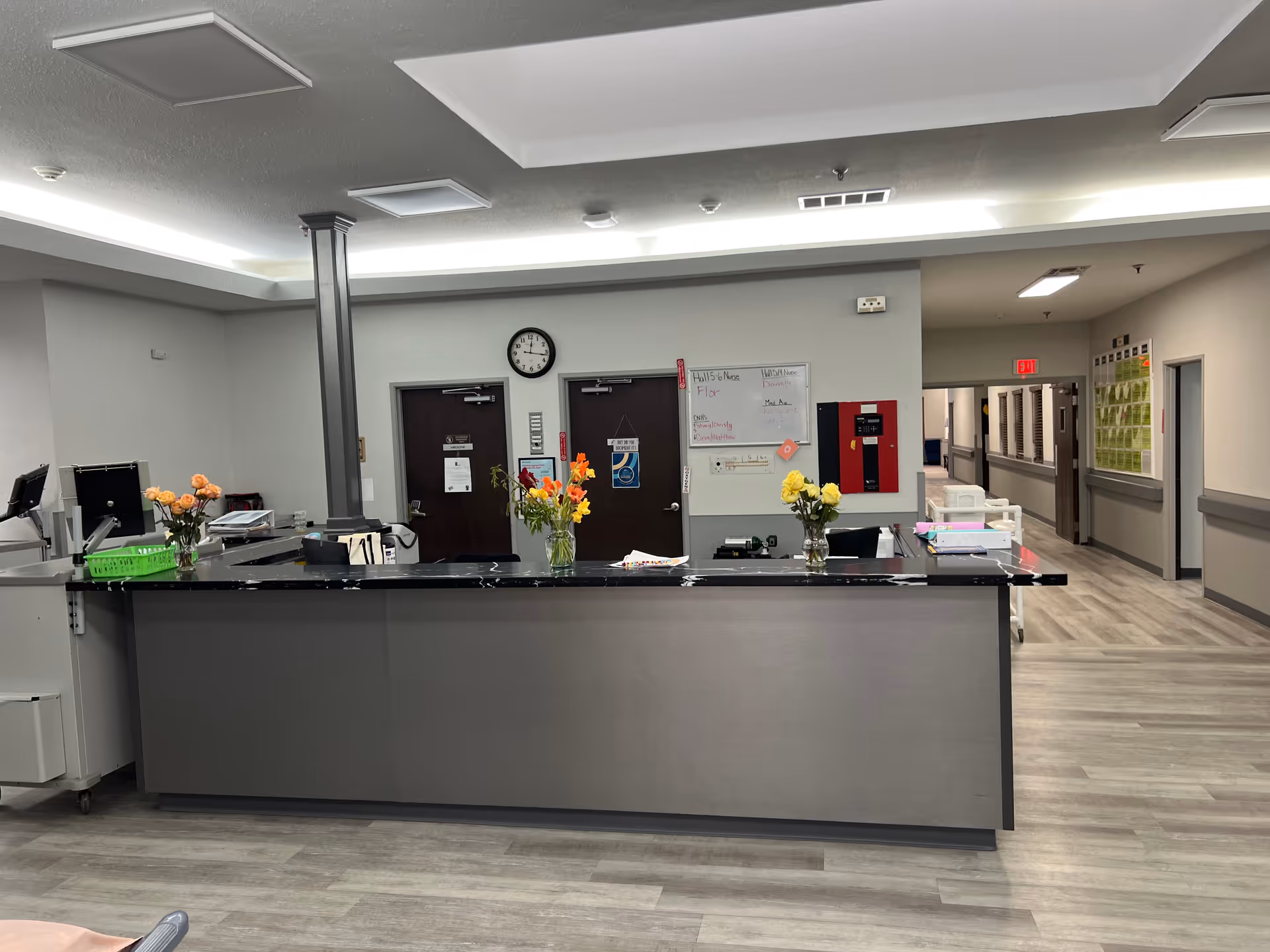 Reception desk area in a nursing and rehabilitation center with a black countertop, vases of flowers, computer monitors, and a hallway leading to other rooms. The walls are light gray with a clock and whiteboard mounted behind the desk.