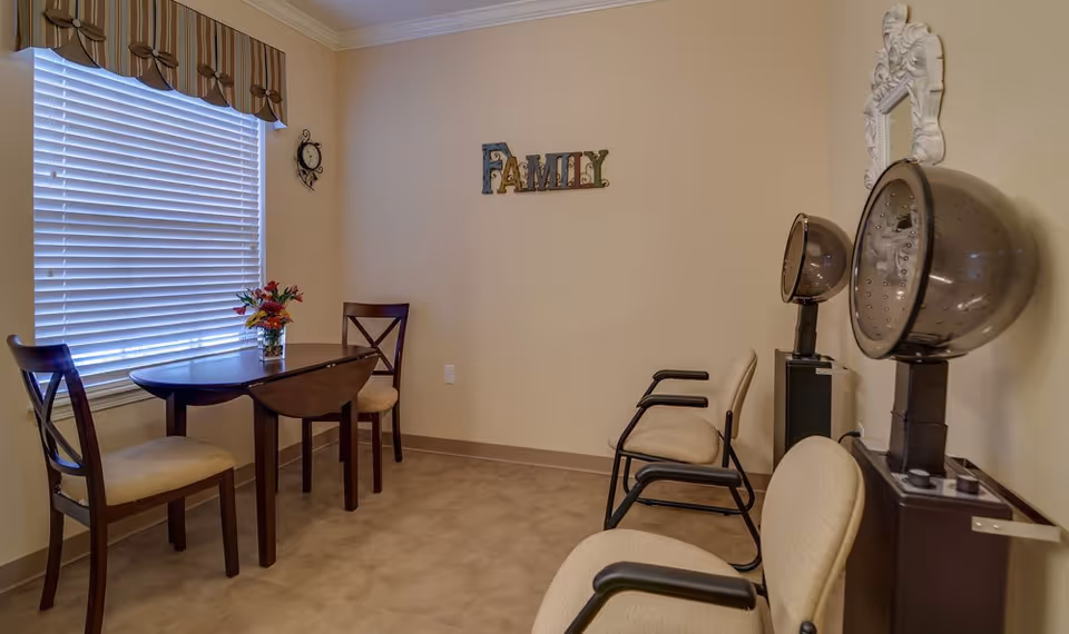 Interior salon-style common room with a small table and chairs by a window and two hooded hair dryers along the wall beneath a 'FAMILY' sign.