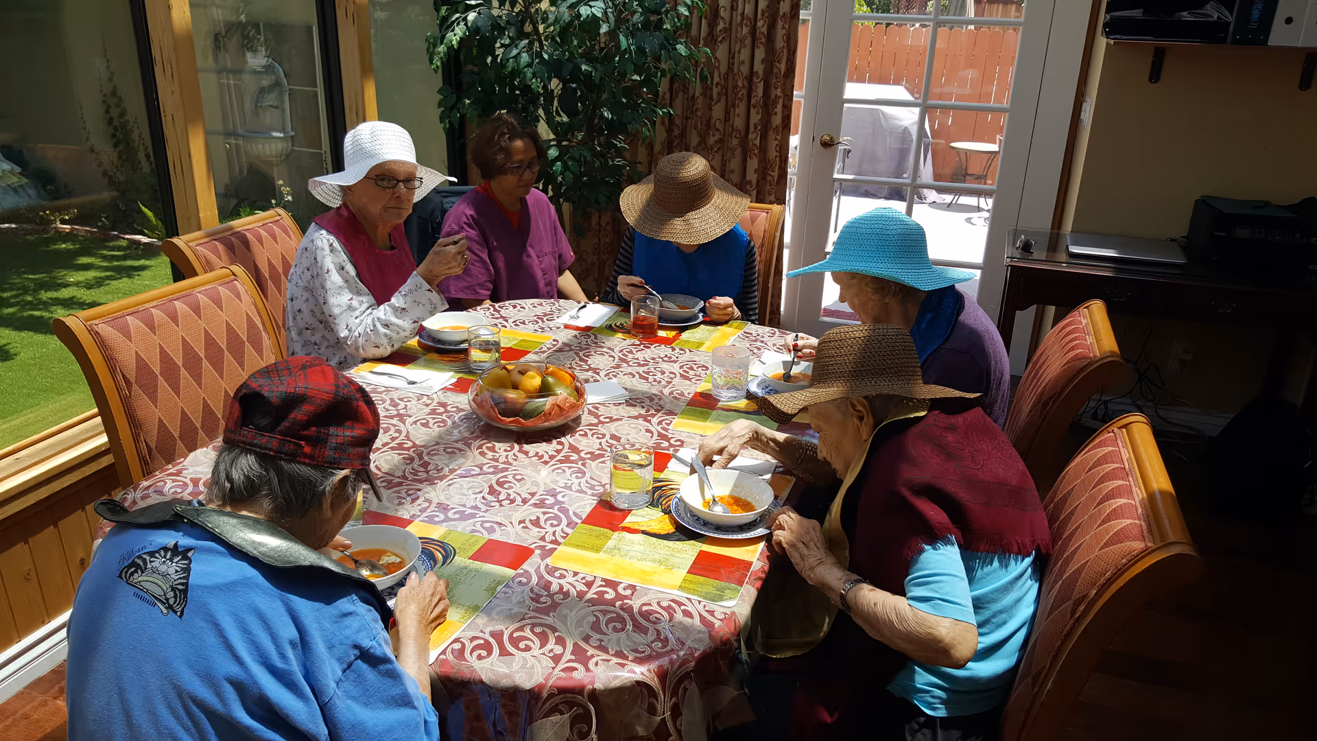 A group of elderly women wearing hats sitting around a dining table inside a room, eating soup. The table is covered with a patterned tablecloth and colorful placemats, with a bowl of fruit in the center. There is a large window showing a green lawn outside and a glass door leading to a patio area.
