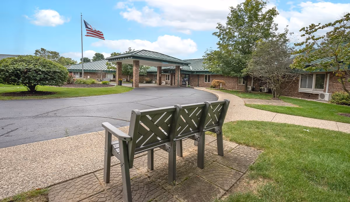 Outdoor view of Valley Pines Senior Living facility showing a paved driveway with a covered entrance, an American flag on a flagpole, a three-seat bench on a paved area, green grass, bushes, and trees under a partly cloudy sky.