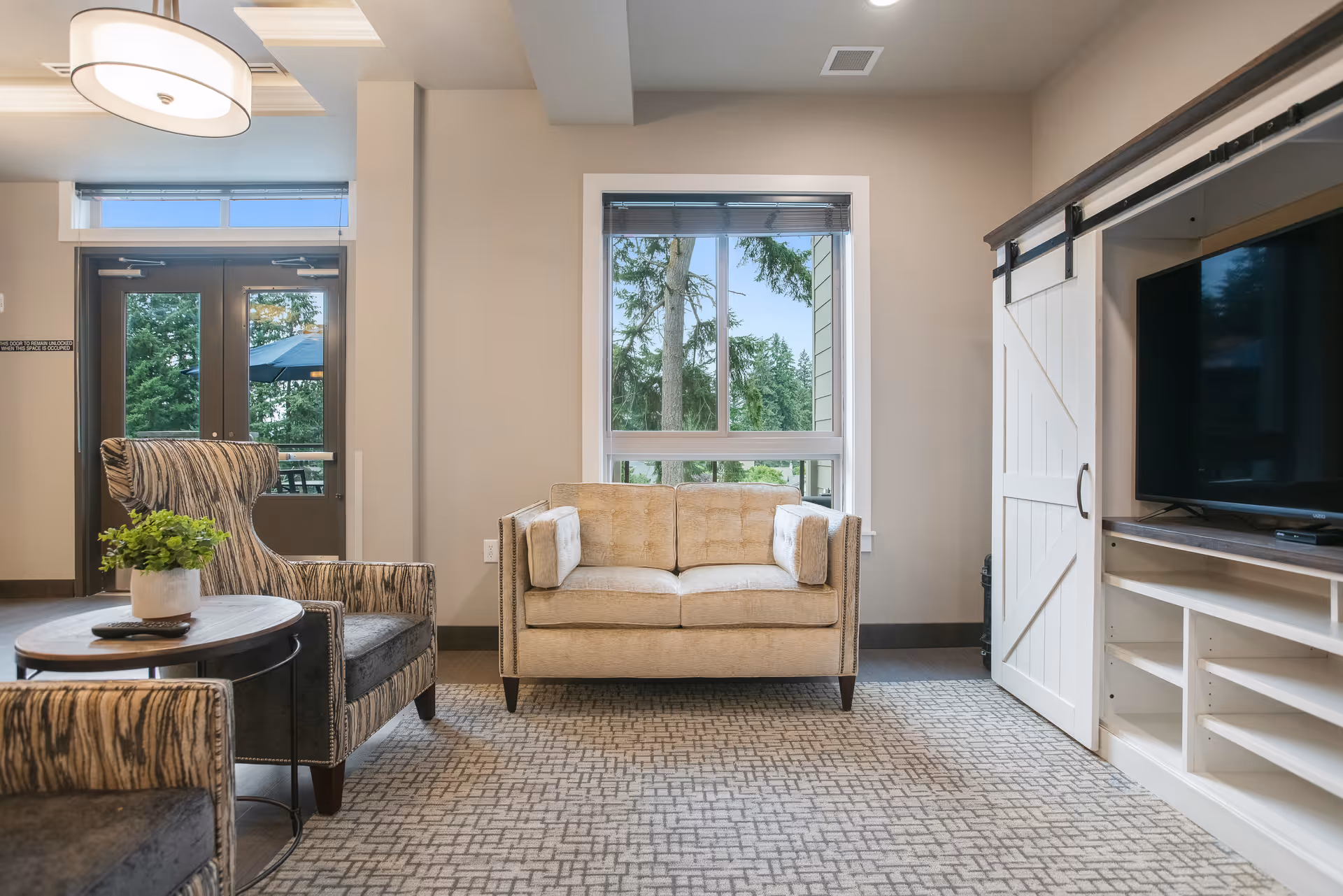 Well-lit living room featuring a beige loveseat centered under a window, patterned armchairs and side tables, and an entertainment cabinet with a sliding barn door.