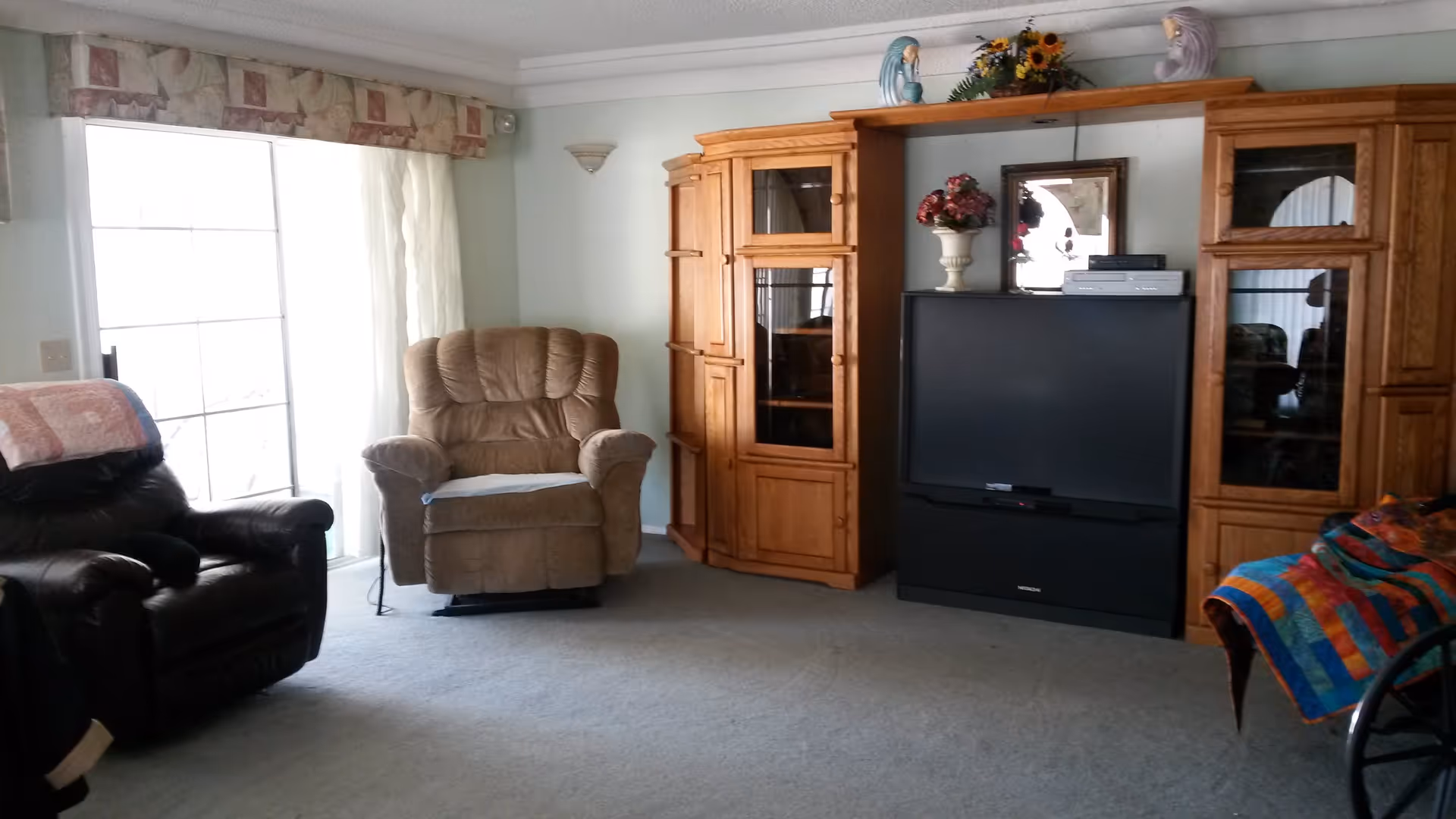 A living room with two recliner chairs, one black and one beige, positioned near a large window with sheer curtains. There is a wooden entertainment center with glass cabinets and a large television in the center. Decorative items including flowers and figurines are placed on top of the entertainment center. A colorful quilt is draped over a piece of furniture on the right side.