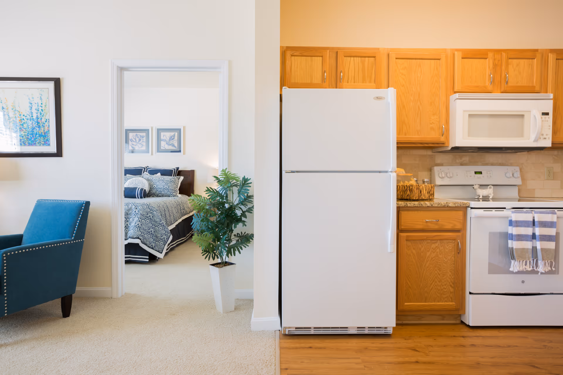 Interior view showing a kitchen area with wooden cabinets, a white refrigerator, stove, and microwave. To the left, there is a doorway leading to a bedroom with a bed covered in blue and white bedding. A blue armchair and a potted plant are also visible in the foreground.