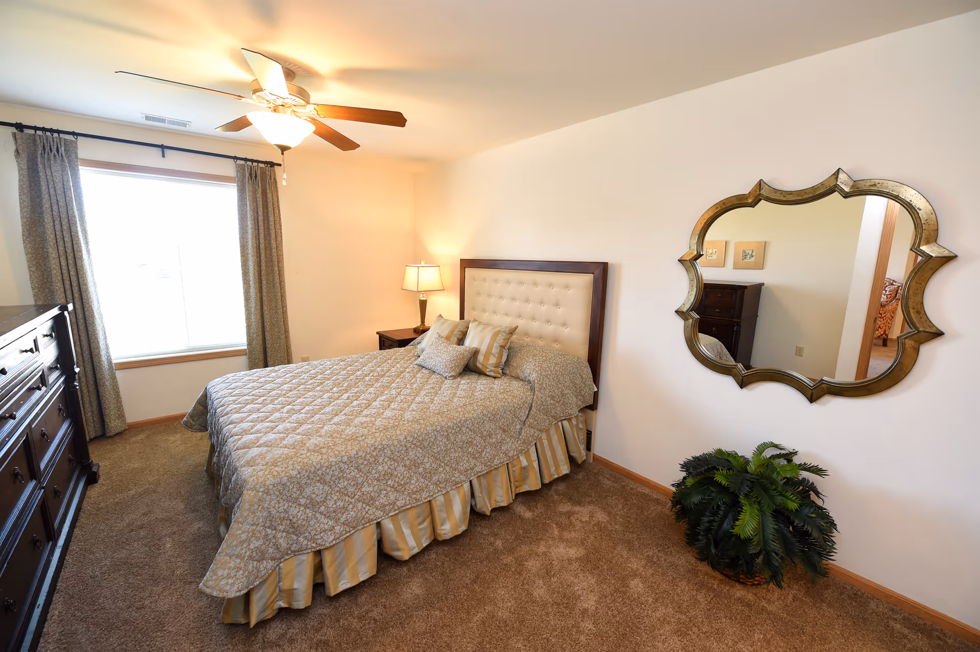 A bedroom with a bed covered in a patterned quilt and pillows, a wooden dresser, a nightstand with a lamp, a ceiling fan with light, a large window with curtains, a decorative wall mirror, and a potted plant on the carpeted floor.