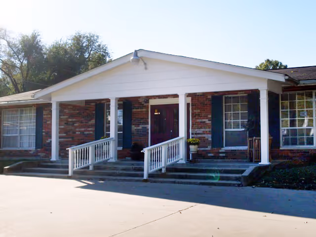 Front exterior view of a single-story brick building with white columns and railings leading up to a covered entrance. The building has large windows with dark shutters and a concrete driveway in front.