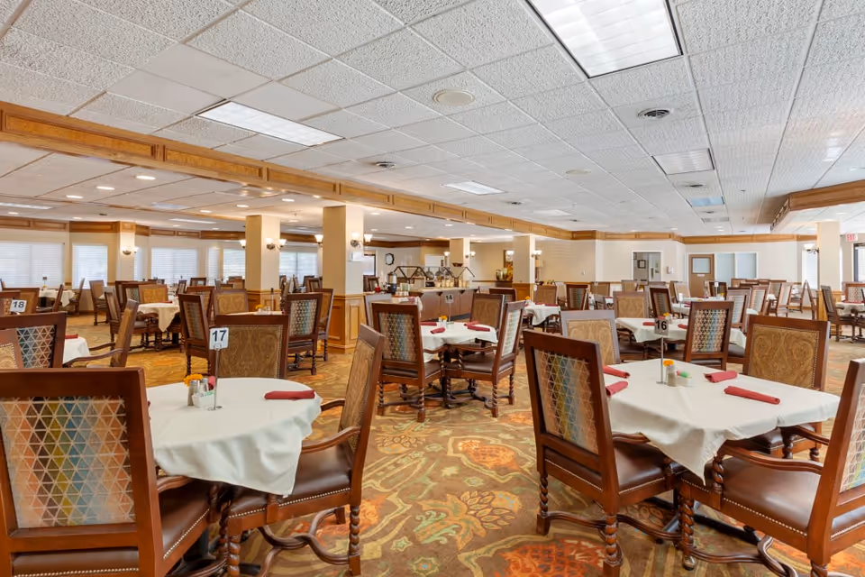Spacious dining room with many round tables covered in white tablecloths, wooden chairs, and numbered place settings in an assisted living facility.
