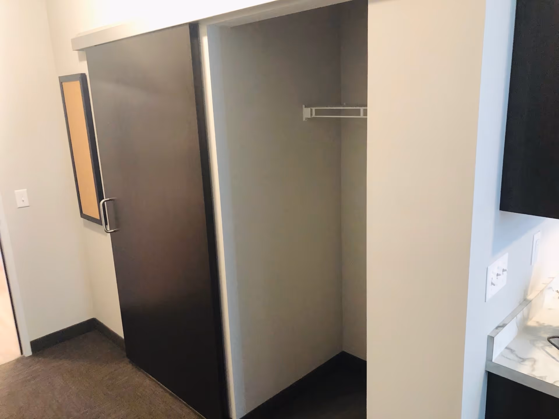 Interior view of a room corner with a sliding dark brown door partially open revealing an empty closet with a single white shelf and hanging rod. The walls are light-colored, and part of a kitchen counter with a marble-like surface and dark cabinetry is visible on the right side.