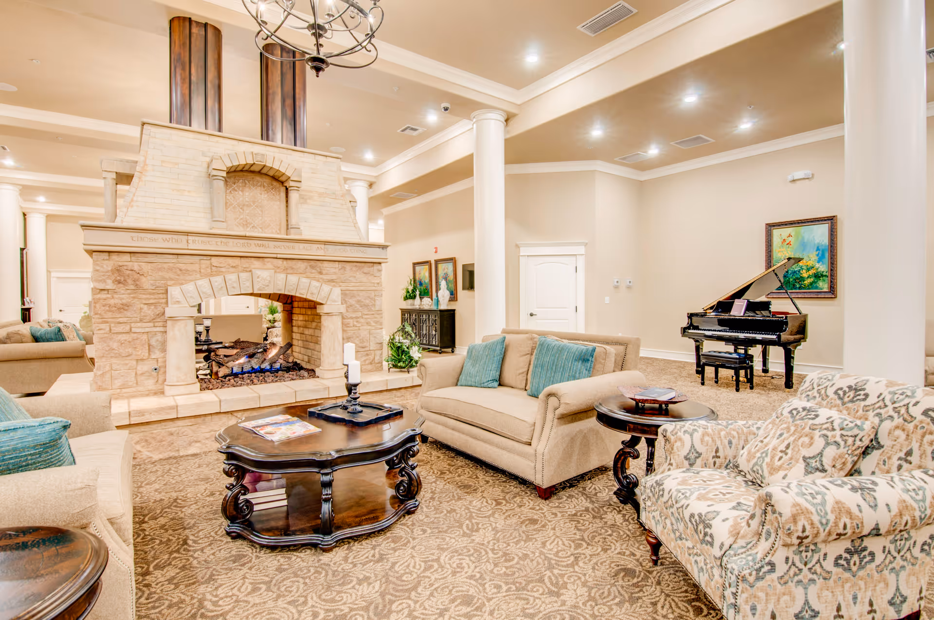 A spacious and elegant living room area with beige walls and carpet. The room features a large stone fireplace with a fire burning, surrounded by comfortable sofas and armchairs with patterned and solid cushions. A dark wooden coffee table and side table are placed near the seating. In the background, there is a black grand piano and a colorful painting on the wall. The ceiling has recessed lighting and a decorative chandelier.