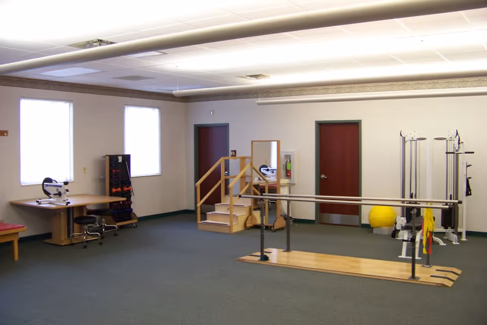 A spacious physical therapy or rehabilitation room with parallel bars on a wooden platform, exercise equipment including a yellow exercise ball, a small set of stairs with handrails, a table with exercise devices, and two windows letting in natural light.