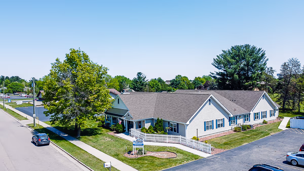 A single-story building with beige siding and a brown roof, surrounded by green grass and trees under a clear blue sky. There is a white picket fence in front of the building and a sign that reads 'Our House Senior Living Wisconsin Rapids Memory Care'. Several cars are parked on the adjacent street and parking lot.