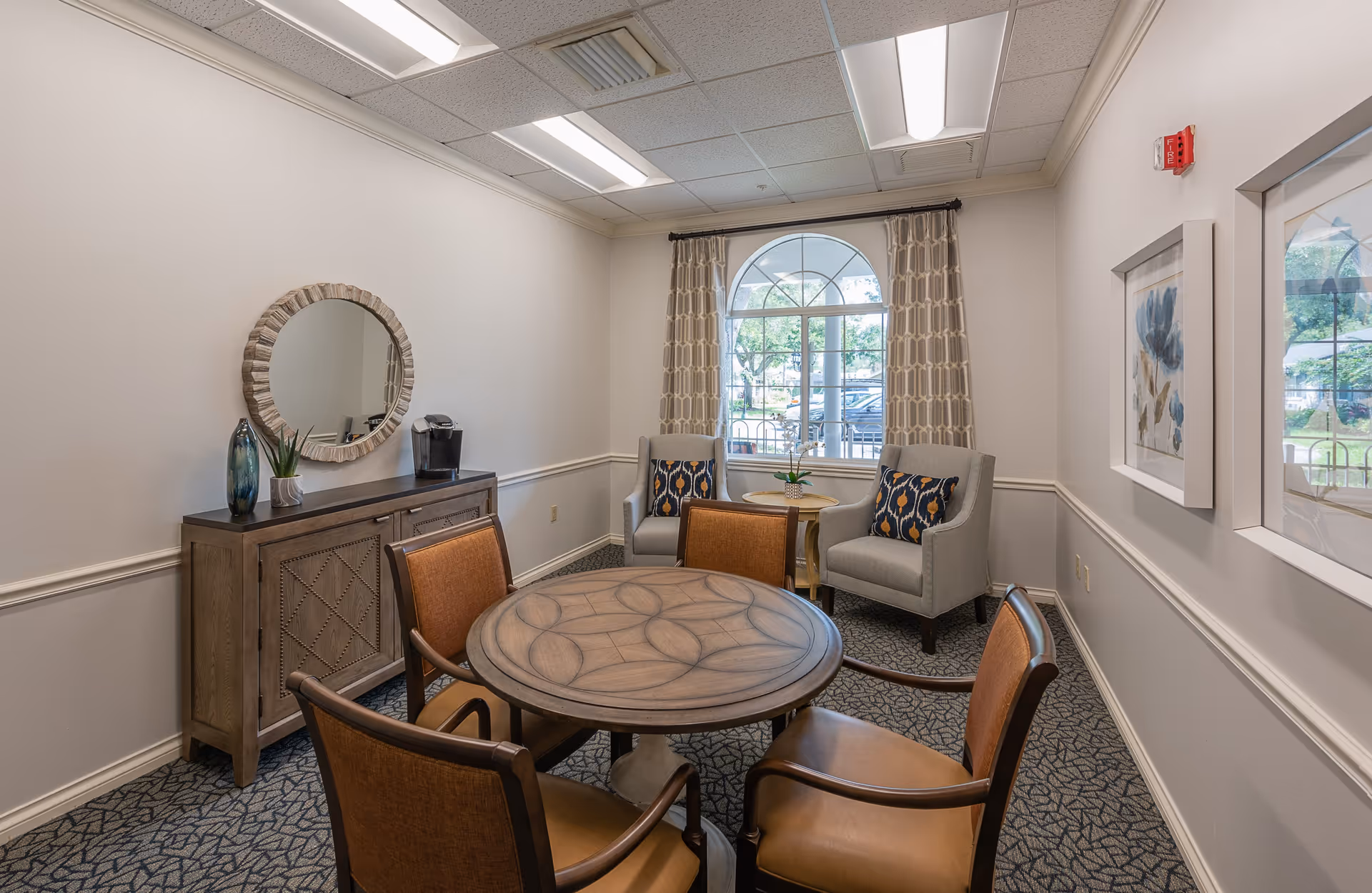 A small sitting area in a senior living facility with a round wooden table surrounded by four brown cushioned chairs. In the background, there are two gray armchairs with patterned pillows next to a window with patterned curtains. A wooden sideboard with a round mirror, a vase, a small plant, and a coffee maker is against the left wall. The walls are light-colored with white trim, and the ceiling has recessed lighting.