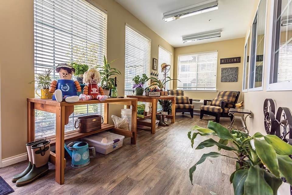 Sunny communal sitting area with wooden tables holding plants and dolls, and plaid armchairs by large windows.