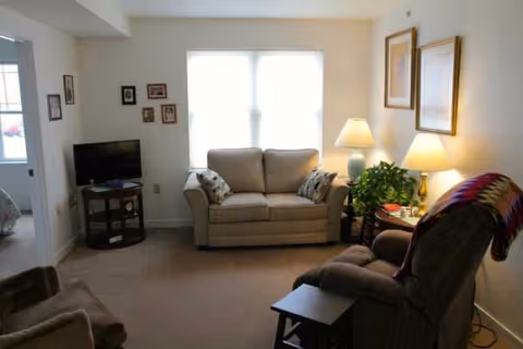 A cozy living room with sofa, chairs, lamps, and a TV in a senior living facility.