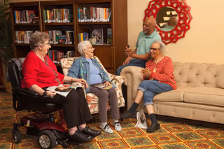 Four elderly people sitting and chatting in a cozy living room area with bookshelves in the background. One woman is in a motorized wheelchair, two women are seated on armchairs, and one man is sitting on a beige sofa. The room has a patterned carpet and a decorative red mirror on the wall.