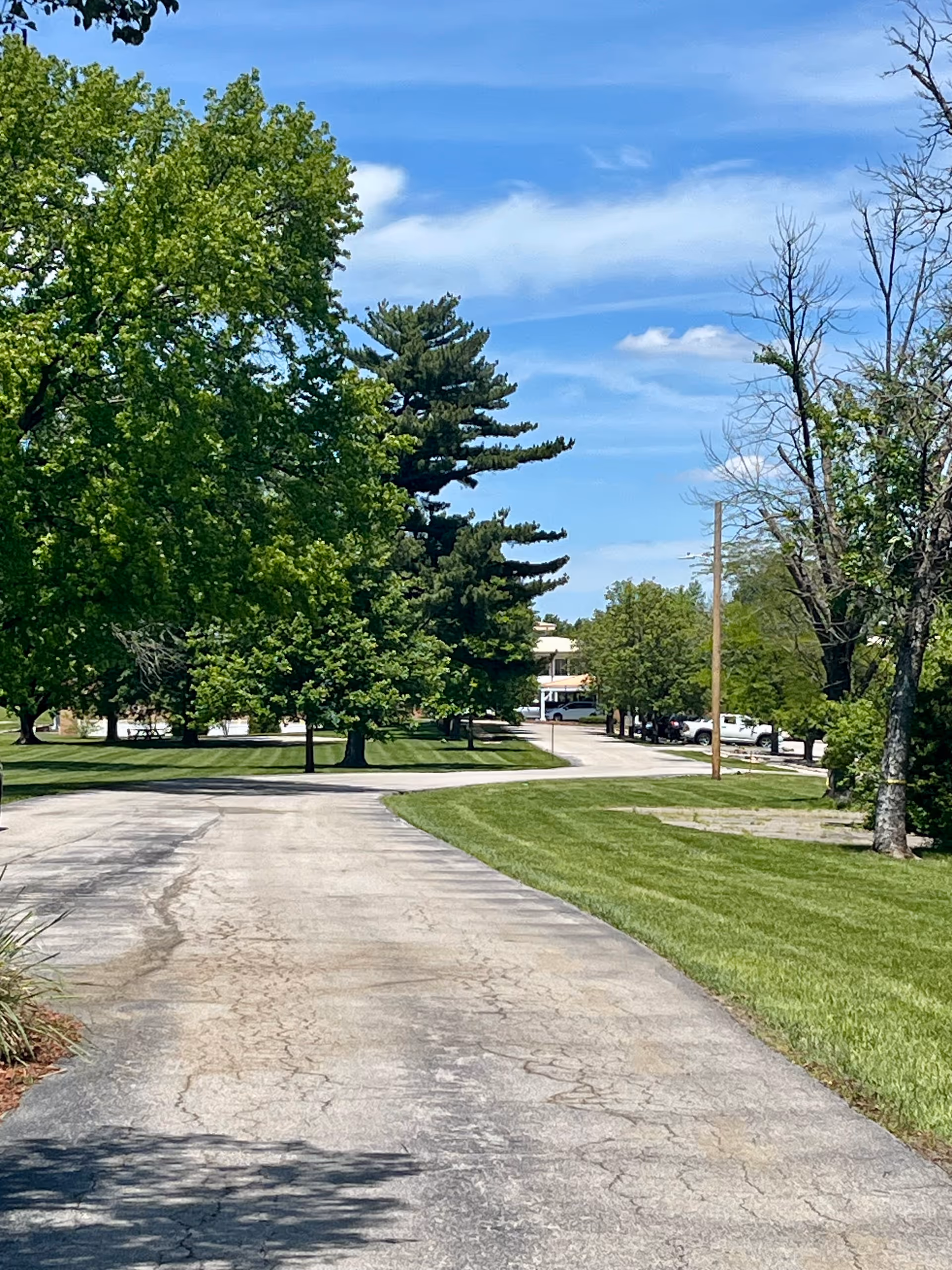 A paved driveway leading towards a building partially visible in the distance, surrounded by green grass and trees under a blue sky with some clouds.