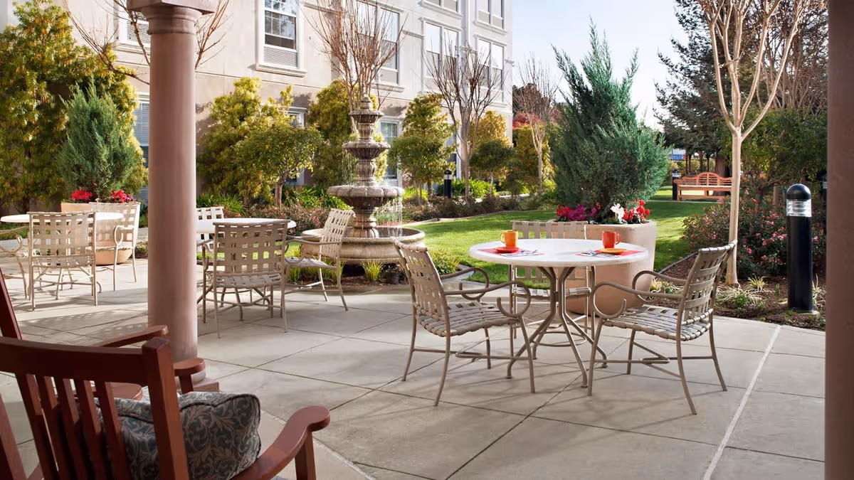 Outdoor patio area at Belmont Village Senior Living Sunnyvale with metal tables and chairs, a stone fountain, potted plants with flowers, green lawn, and trees surrounding the space. The patio is adjacent to the building and has a covered section with wooden rocking chairs and a cushion.