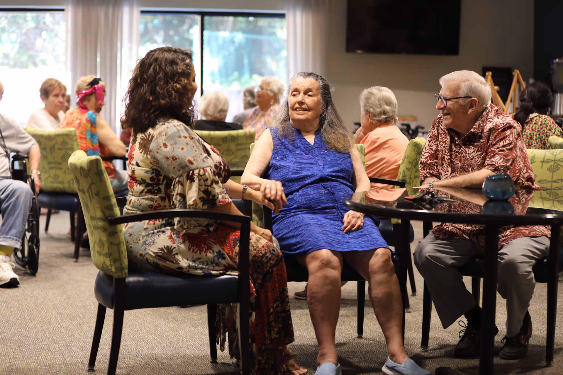 A group of elderly people sitting and socializing in a well-lit common area with green patterned chairs and large windows in the background. Three individuals are seated around a small table, engaged in conversation and smiling.