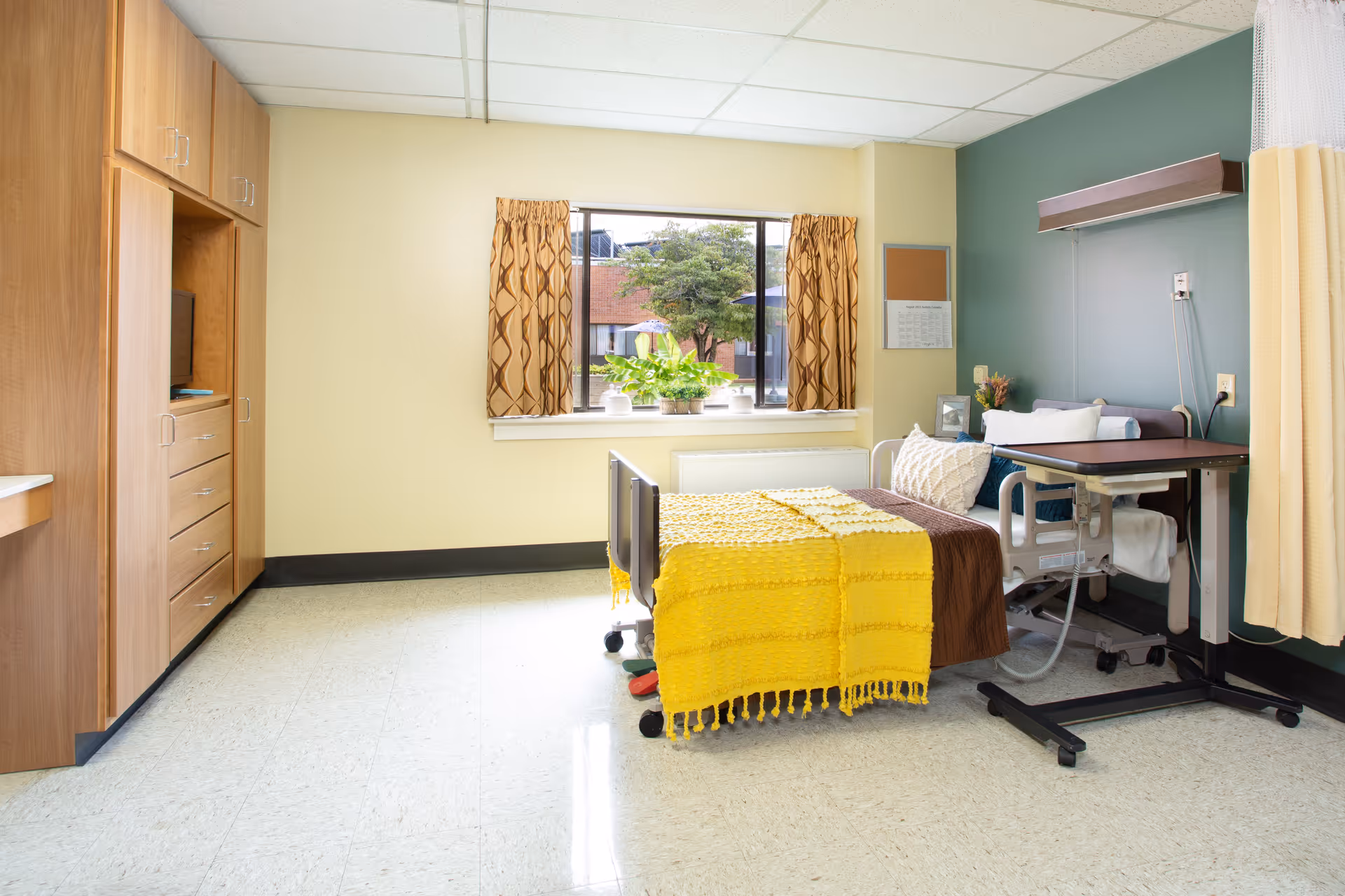 A bright, well-furnished bedroom in a senior living facility with a bed, window, and wooden storage.