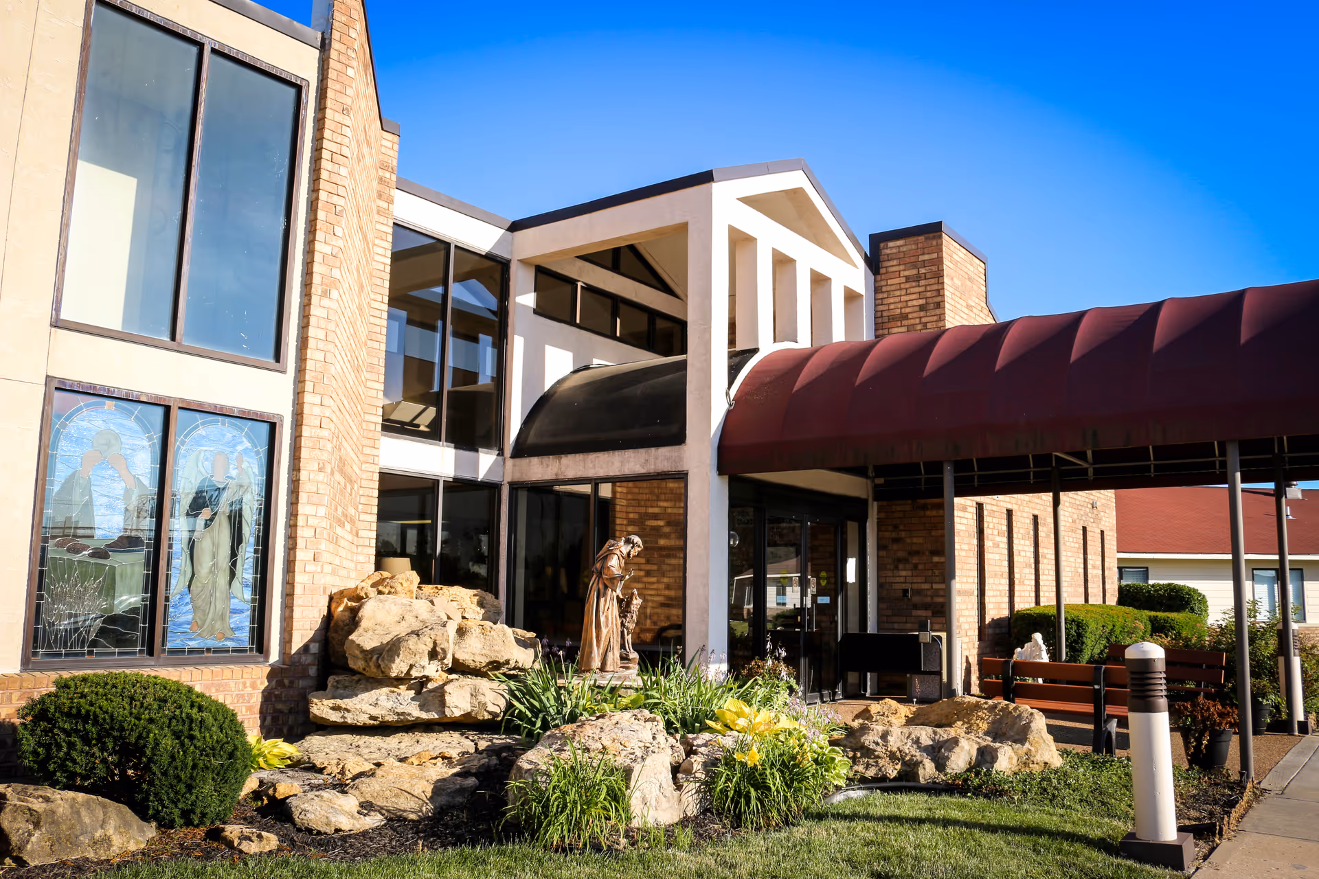 Exterior view of Villa St. Francis Catholic Care Center showing a brick building with large windows, stained glass artwork, a statue, and a covered entrance with a red awning. There are rocks and landscaping with bushes and flowers in front of the building under a clear blue sky.
