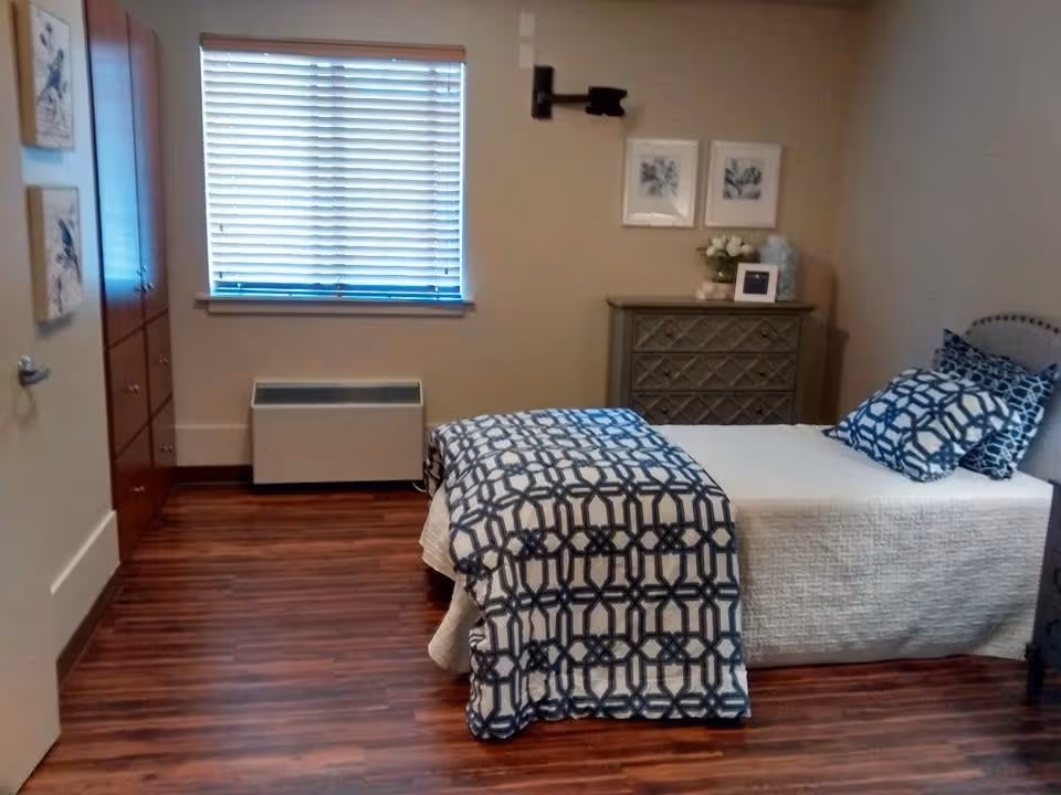 A neatly arranged bedroom with a single bed covered in a white quilt and a blue patterned blanket and pillows. There is a wooden dresser with decorative items and framed pictures on the wall above it. The room has wooden flooring, a window with blinds, and a closed door with two framed artworks on the wall beside it.