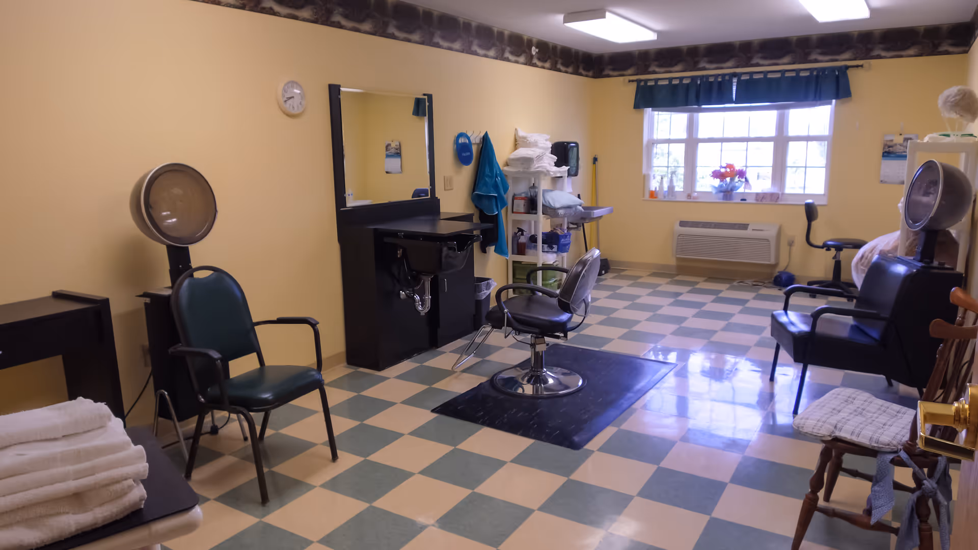 Interior of a hair salon room in a senior living facility with yellow walls and checkered floor tiles. The room contains salon chairs, hair dryers, a large mirror above a black sink station, shelves with towels and supplies, a window with blue curtains, and various chairs arranged around the room.