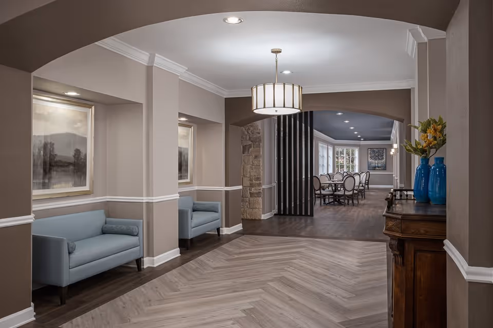 Interior view of a senior living facility hallway with two light blue armchairs placed in recessed wall niches, each accompanied by a framed landscape painting above. The hallway leads to a dining area with a long table and chairs, illuminated by ceiling lights and natural light from large windows. A wooden sideboard with blue vases and yellow flowers is positioned on the right side of the hallway. The walls are painted in neutral tones with white trim, and the flooring transitions from a light herringbone pattern to darker wood in the dining area.