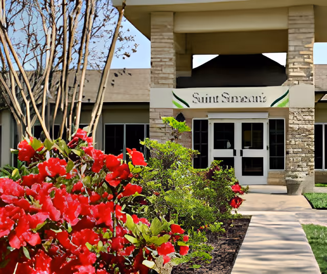 Front entrance of a senior living building with red flowering shrubs and a paved walkway.