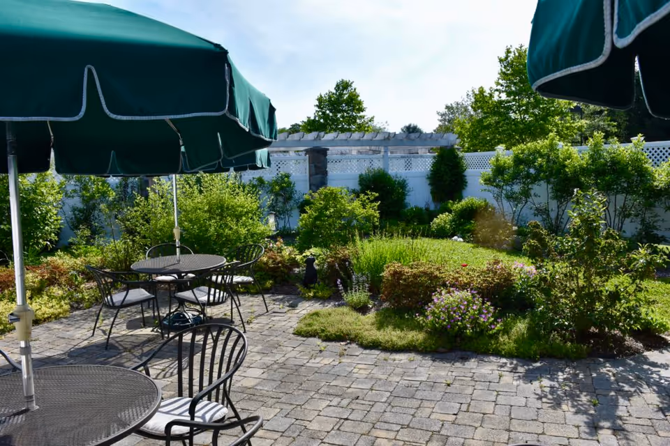 Sunlit courtyard patio with metal tables and green umbrellas overlooking a landscaped garden and white fence.