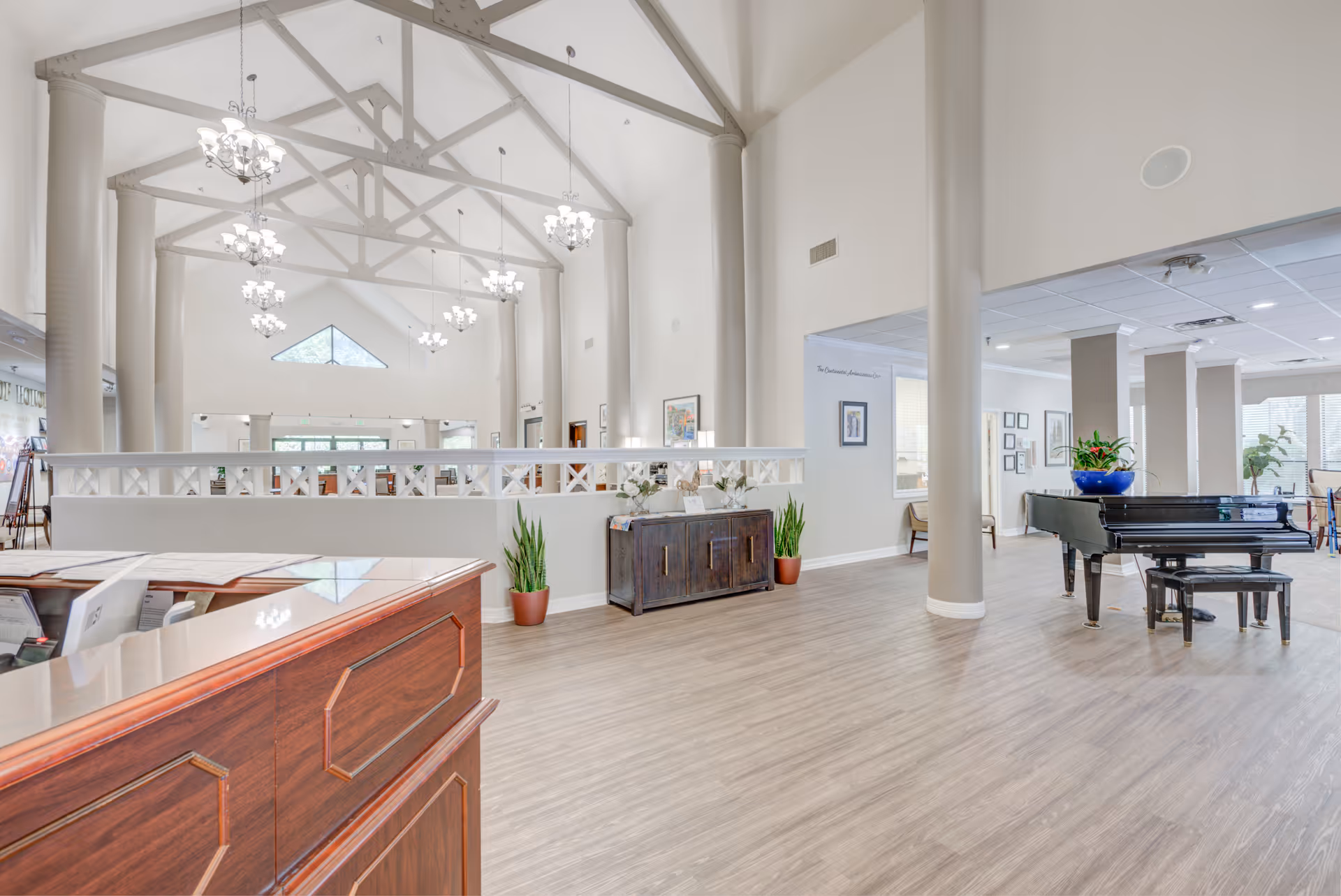 Spacious and bright interior of The Continental Retirement Community featuring a high vaulted ceiling with exposed beams and multiple chandeliers. The room includes a wooden reception desk in the foreground, a black grand piano with a plant on top to the right, and a wooden cabinet with decorative items and plants along the wall. Large windows and light-colored walls enhance the airy atmosphere.