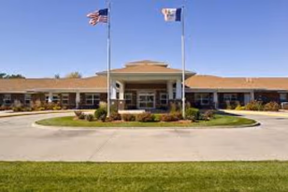 Front exterior view of Addington Place of Des Moines building with two flagpoles displaying the American flag and another flag, a circular driveway, and landscaped greenery under a clear blue sky.