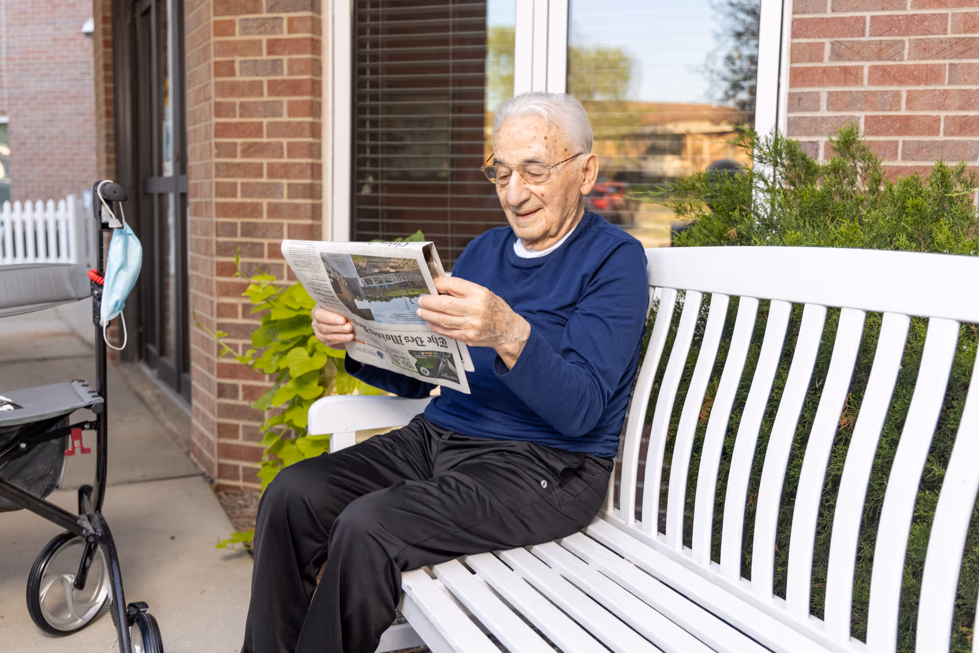 An elderly man with glasses is sitting on a white bench outside a brick building, reading a newspaper. A walker with a face mask hanging on it is positioned nearby. Green plants and a window with blinds are visible behind him.