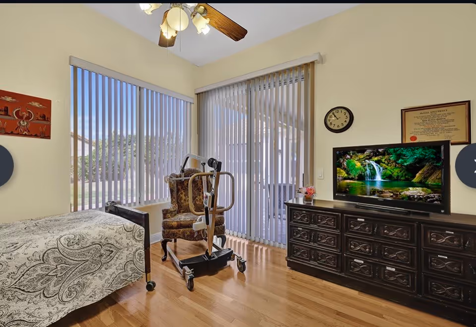 A well-lit bedroom with a single bed covered in a patterned bedspread, a floral upholstered armchair with a patient lift device next to it, a large dark wooden dresser with a flat-screen TV displaying a waterfall scene, a wall clock, a framed certificate, and vertical blinds covering large windows and a sliding glass door.