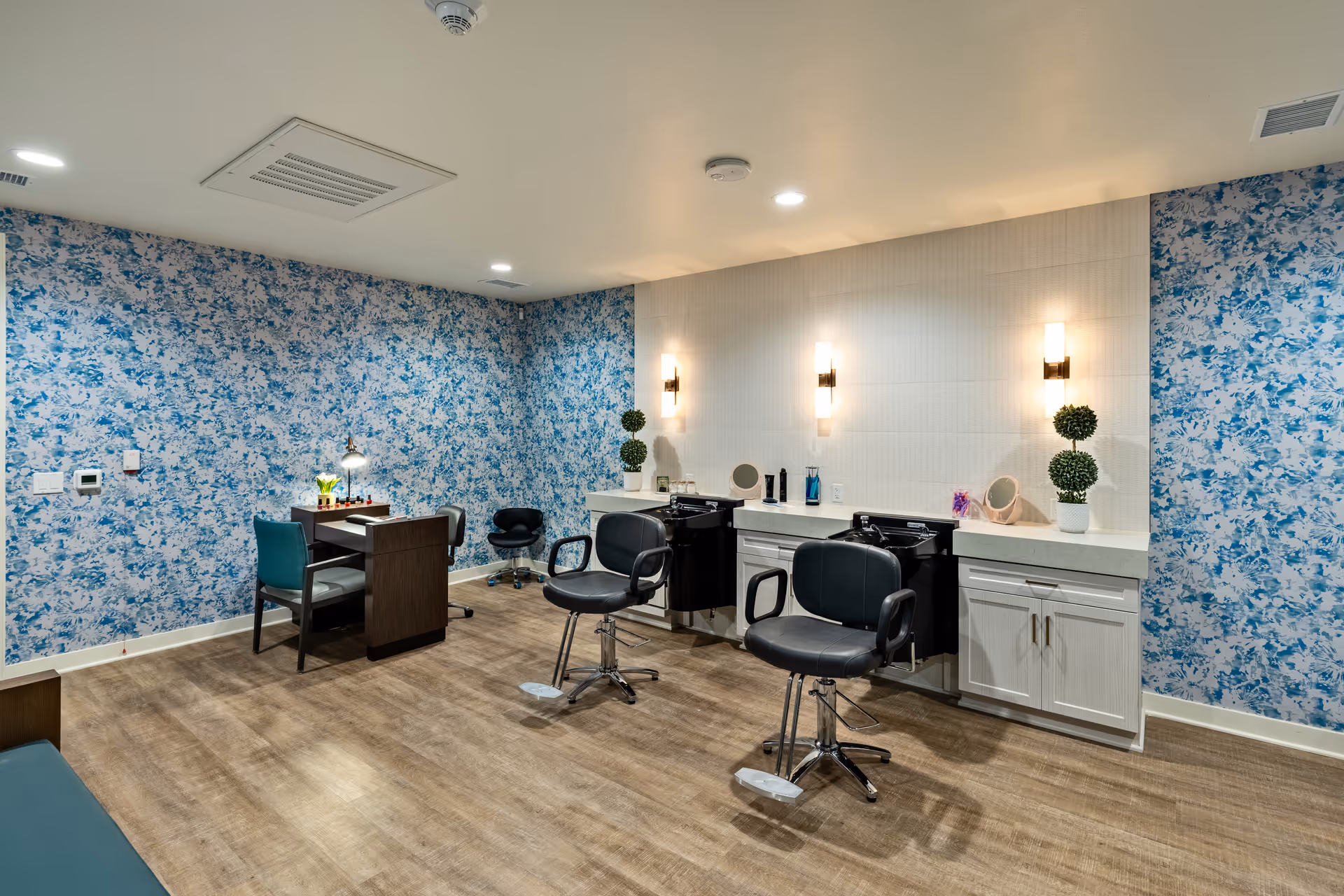 Interior of a salon area with two black salon chairs in front of white counters with sinks, illuminated by wall-mounted lights. The room has blue floral wallpaper on two walls, wood flooring, and a small table with chairs and manicure supplies in the corner.