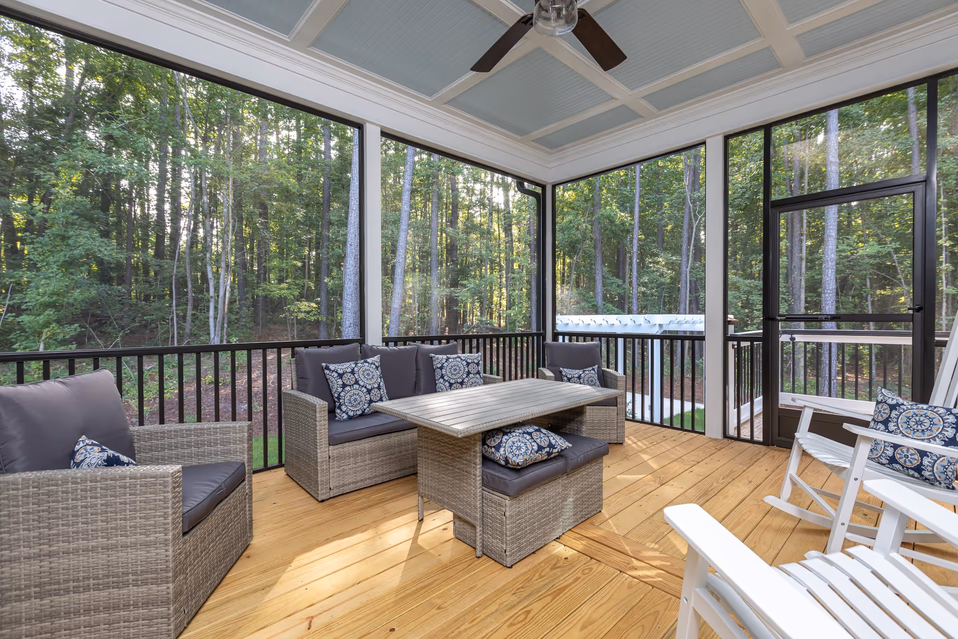 A screened-in porch with wooden flooring and a ceiling fan. The porch is furnished with gray wicker seating including a loveseat, two armchairs, and a bench around a rectangular table. There are decorative blue and white patterned pillows on the seating. The porch overlooks a wooded area with tall trees.