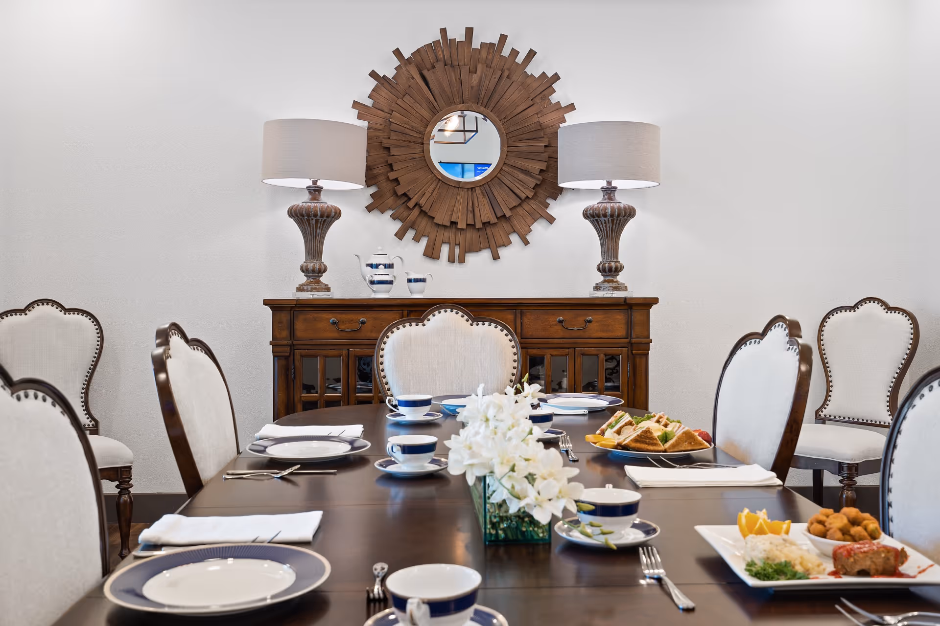 A formal dining room with a dark wooden table set with plates, cups, and silverware. There are upholstered chairs with wooden frames around the table. On the table are plates of food including sandwiches and a meal with vegetables and meat. In the background, there is a wooden sideboard with two matching lamps and a decorative round wooden mirror on the wall.