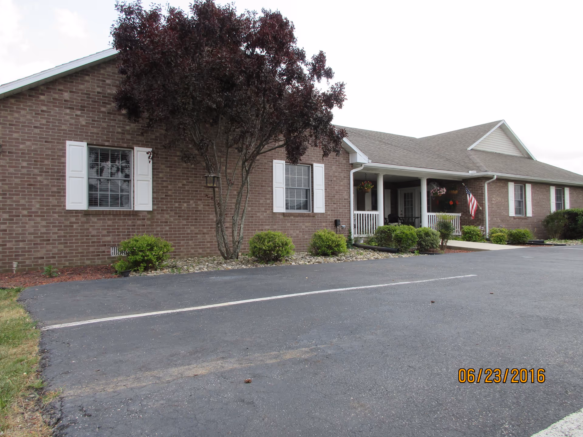Exterior view of a single-story brick building with white window shutters and a covered porch with white railings. There are shrubs and a tree in front of the building, and an American flag is displayed near the porch. The foreground shows a paved parking area with marked parking spaces.