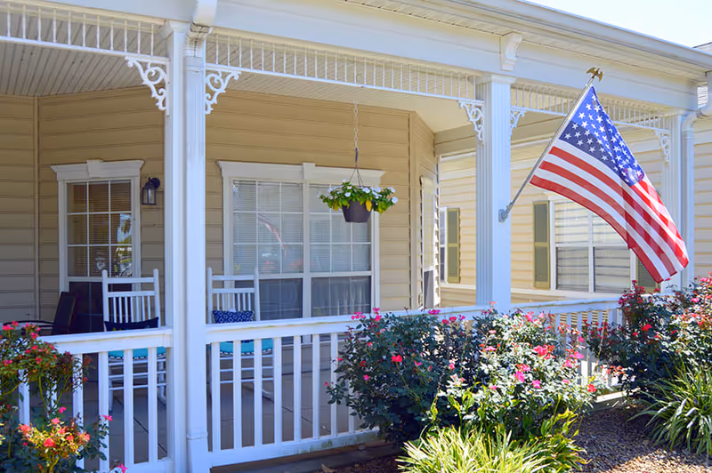 Front porch of a beige senior living facility with white railings and columns, two white rocking chairs with cushions, a hanging flower pot, blooming bushes, and an American flag mounted on the porch.